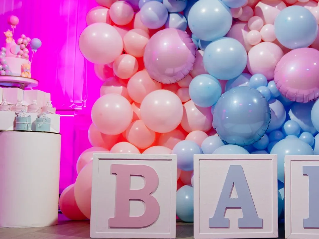 Pink and blue baby shower display with a large balloon wall, pastel letter blocks and a cake on a white pedestal.