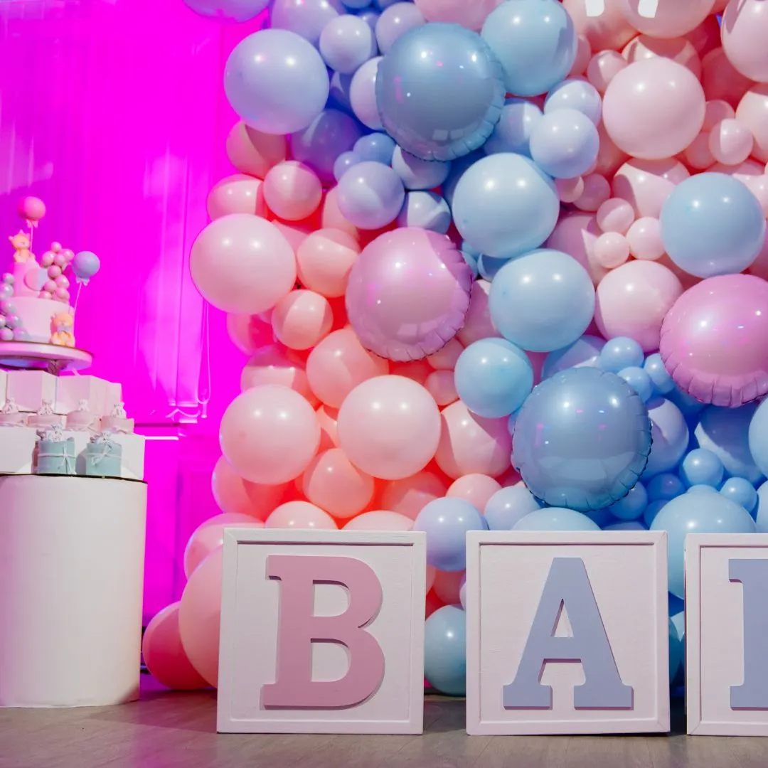 Pink and blue baby shower display with a large balloon wall, pastel letter blocks and a cake on a white pedestal.