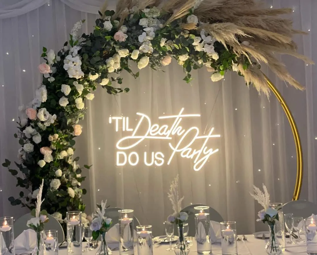 Elegant wedding reception backdrop with a gold circular arch, white and blush flowers, pampas grass and a glowing 'Til Death Do Us Party' neon sign.