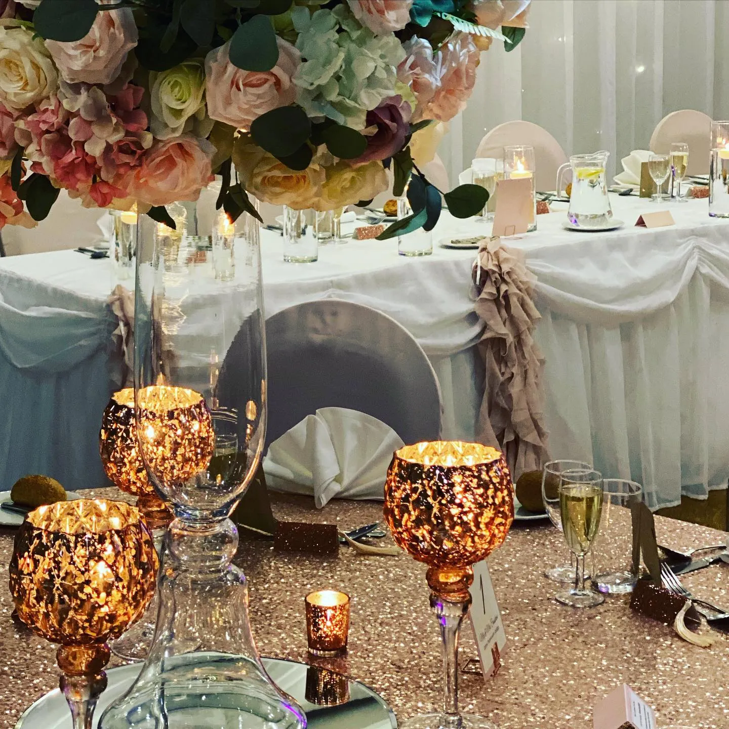Elegant wedding reception table with tall pastel floral centrepiece, glowing candle holders and sparkling rose gold table linen
