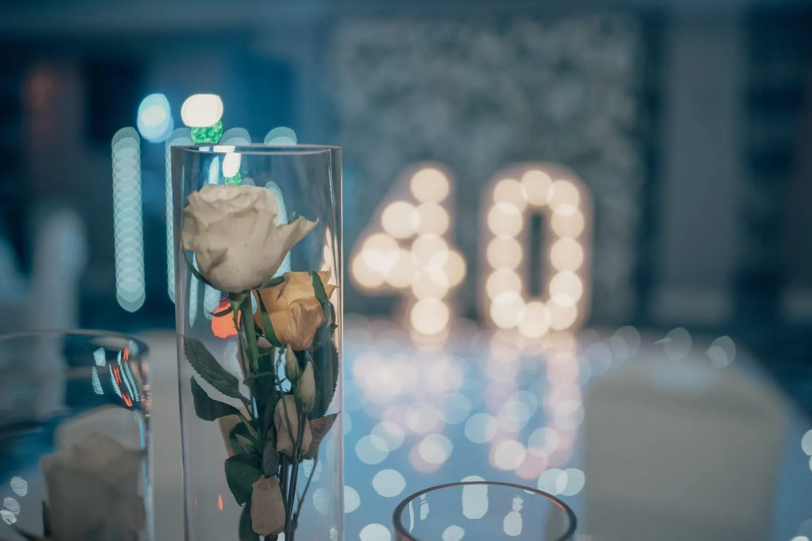 White and cream roses in a tall glass vase on a decorated table, with illuminated number 40 lights softly blurred in the background.