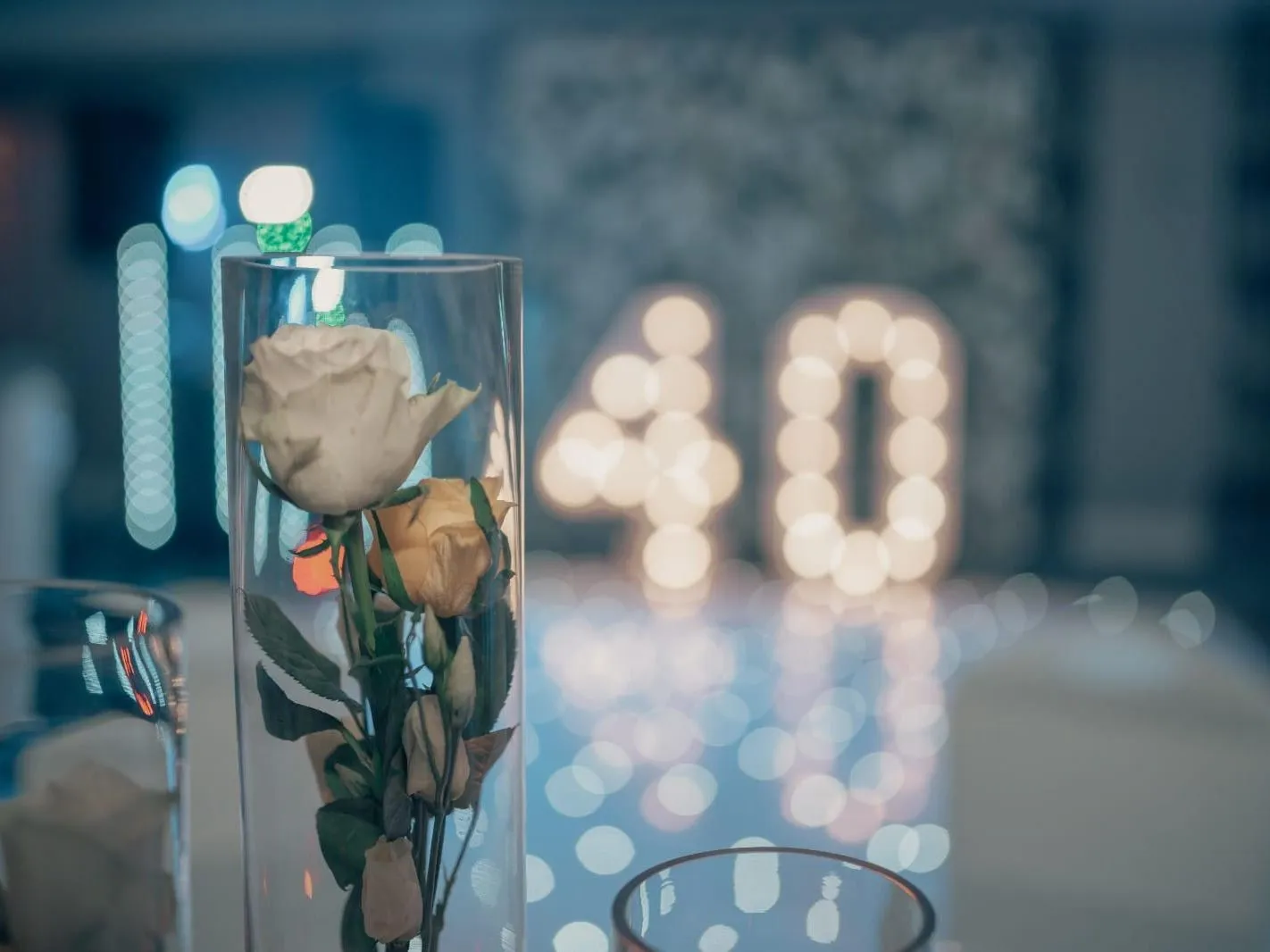 White and cream roses in a tall glass vase on a decorated table, with illuminated number 40 lights softly blurred in the background.