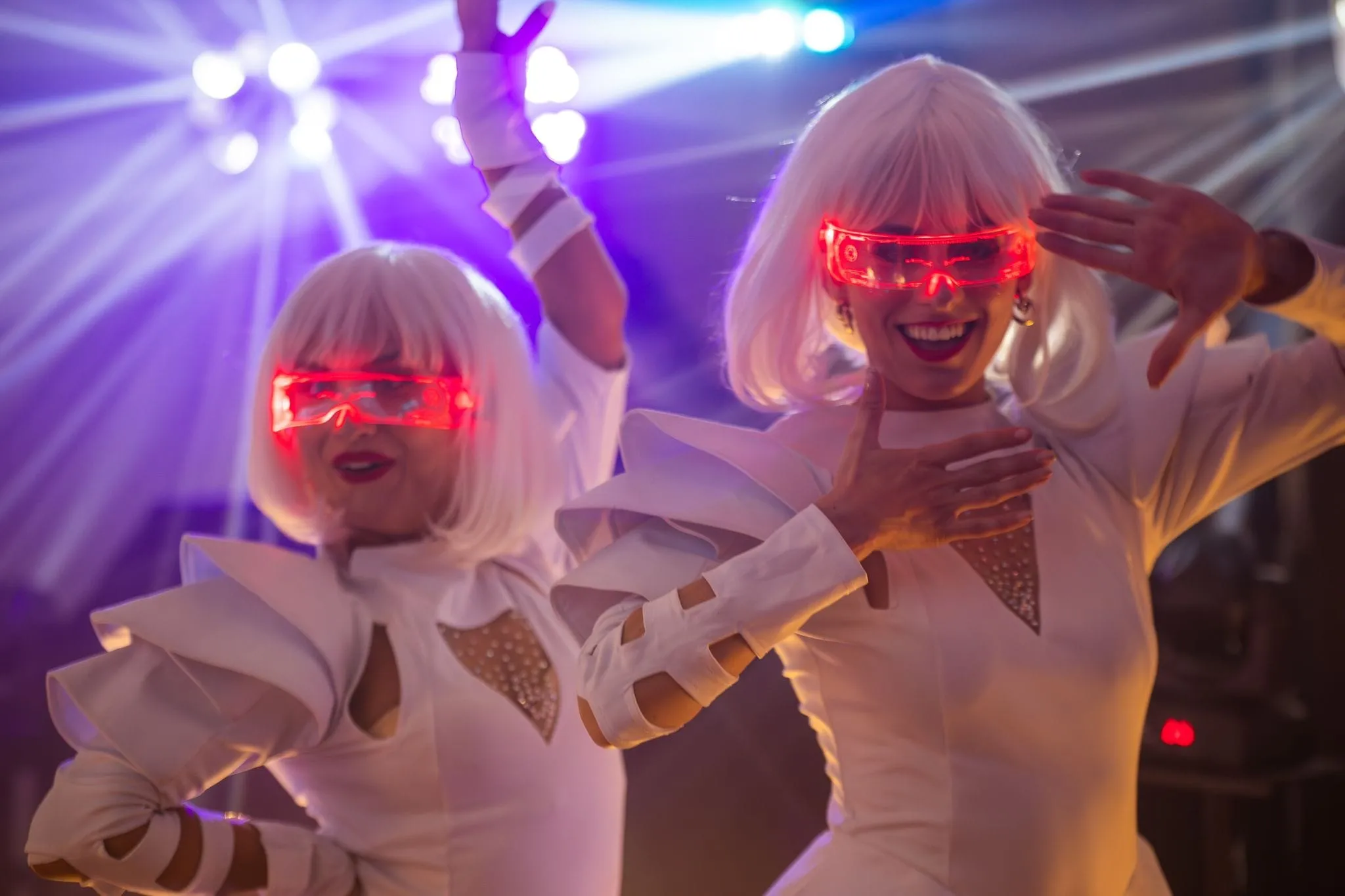 Two performers in white futuristic costumes and glowing red visor glasses dancing under bright stage lights.