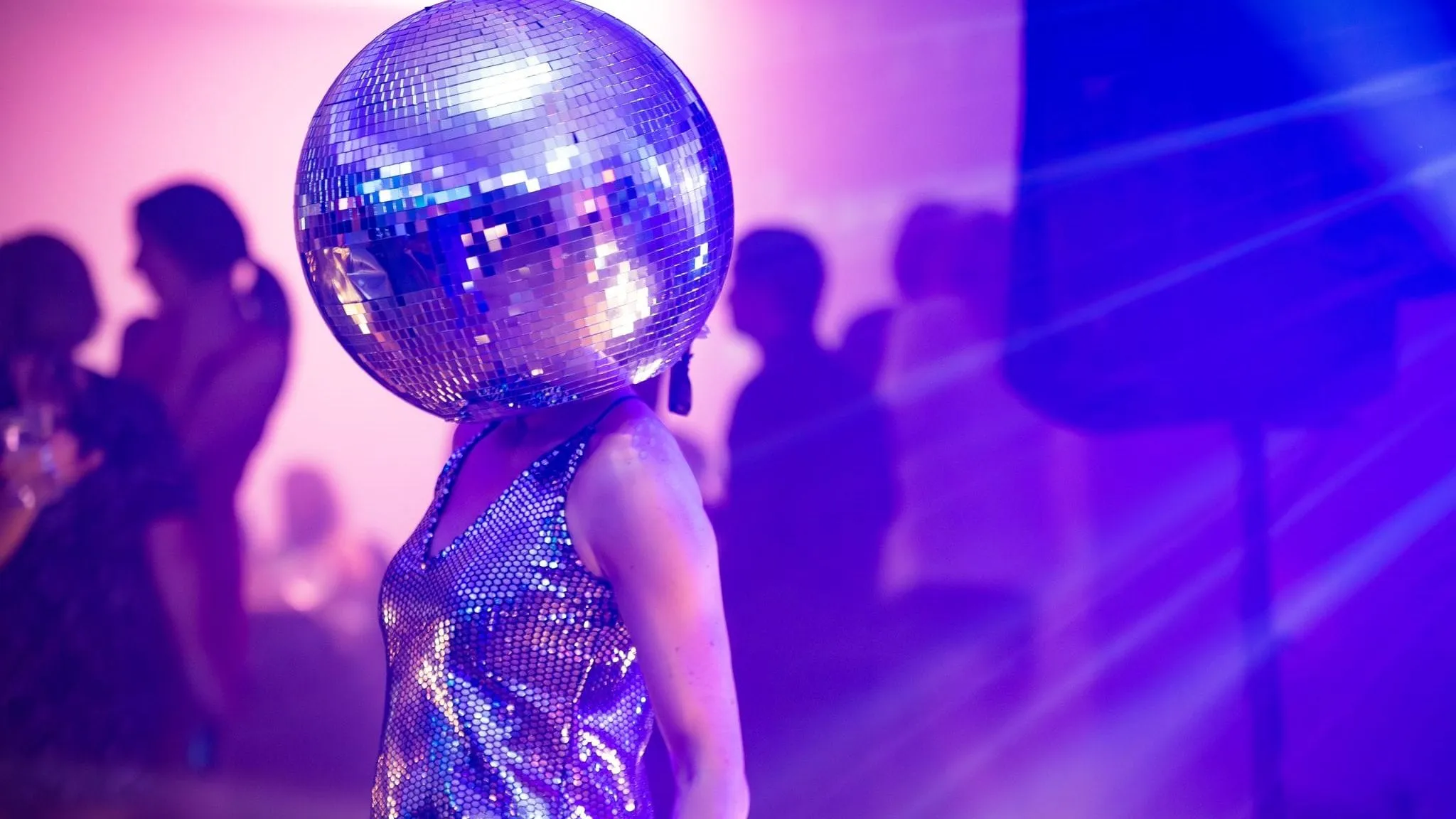 Person wearing a mirrored disco ball helmet and a shimmering silver dress at a party with purple lighting.