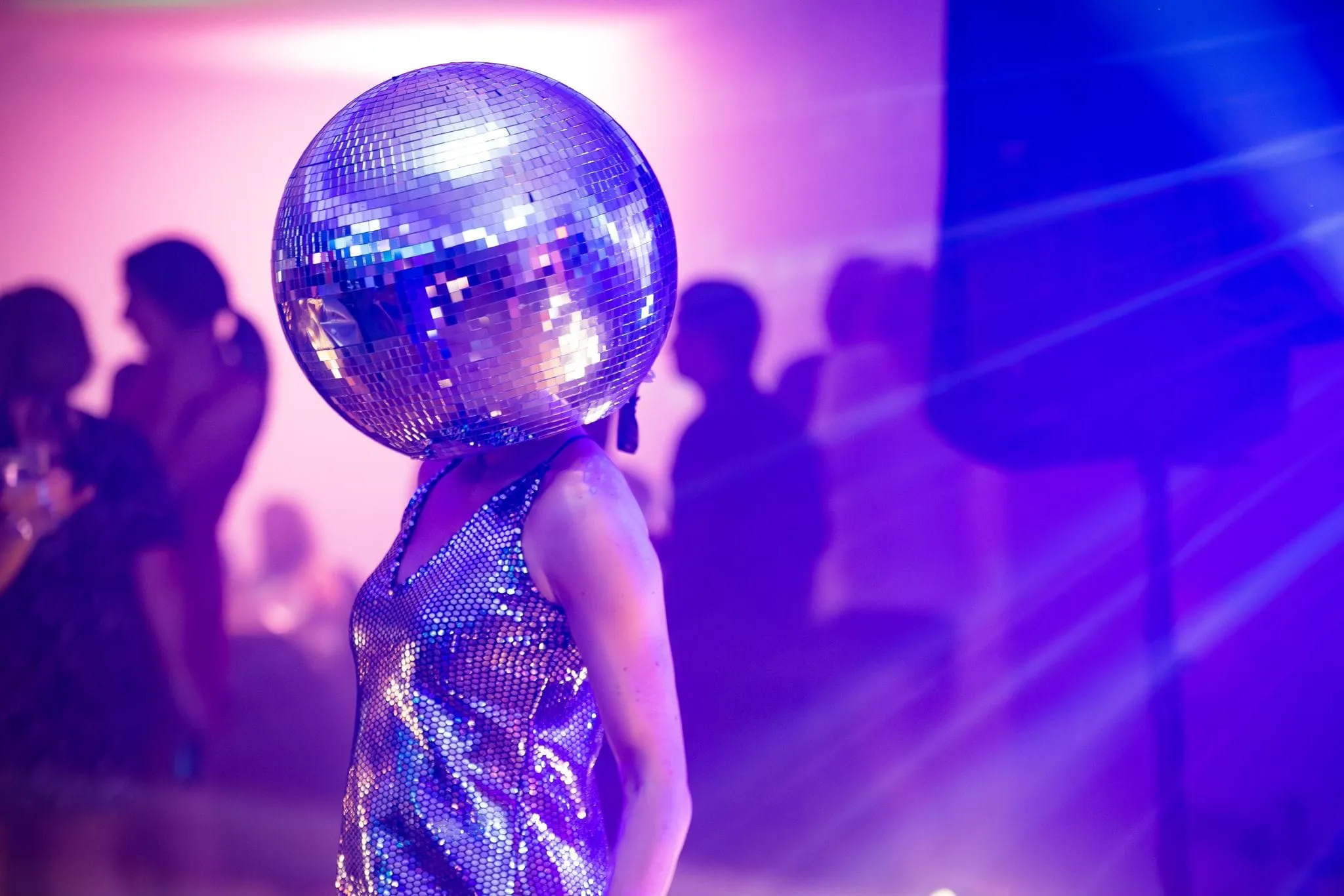 Person wearing a mirrored disco ball helmet and a shimmering silver dress at a party with purple lighting.