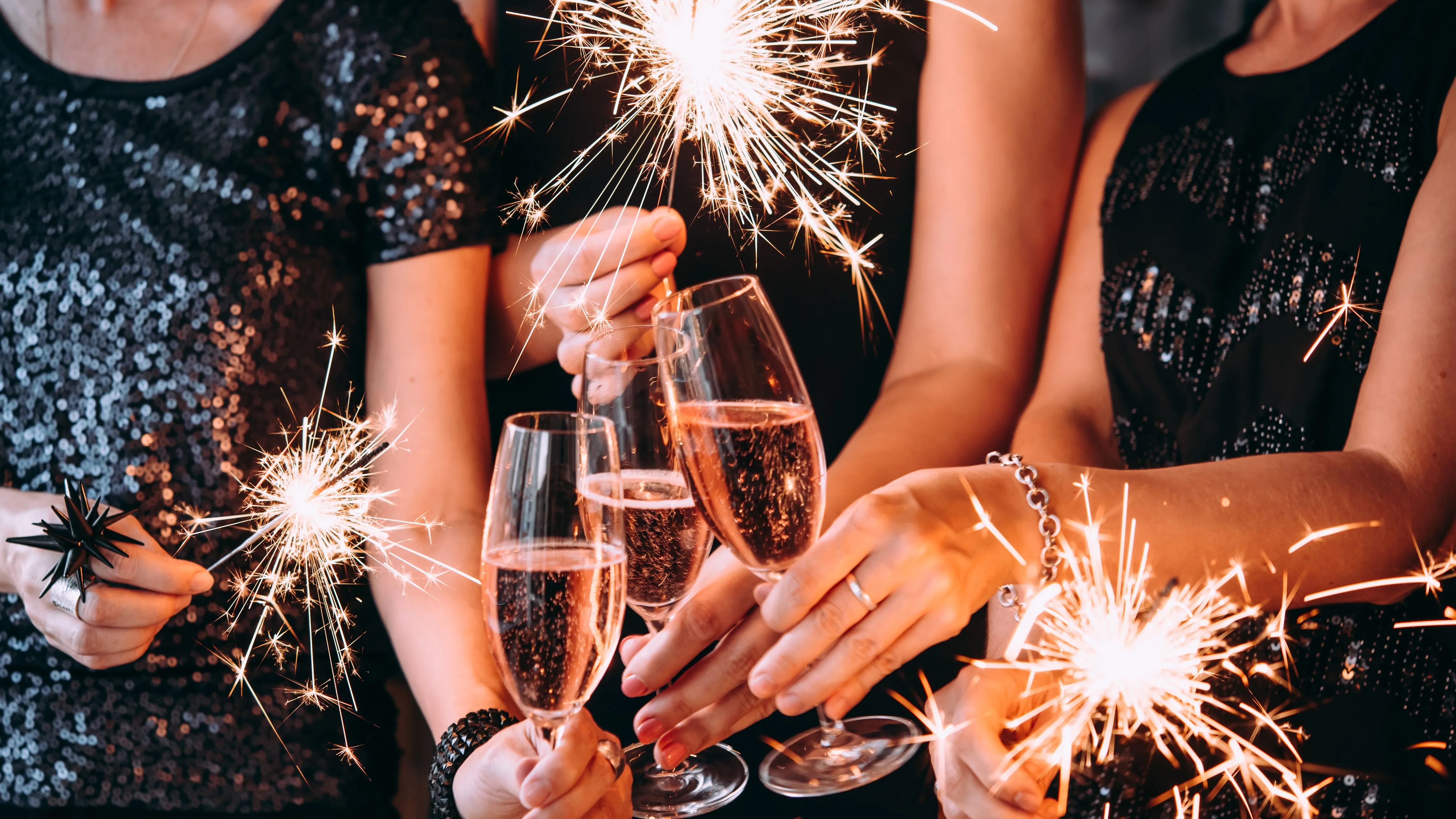 Three women in sequinned black dresses holding sparklers and champagne flutes at a celebration.