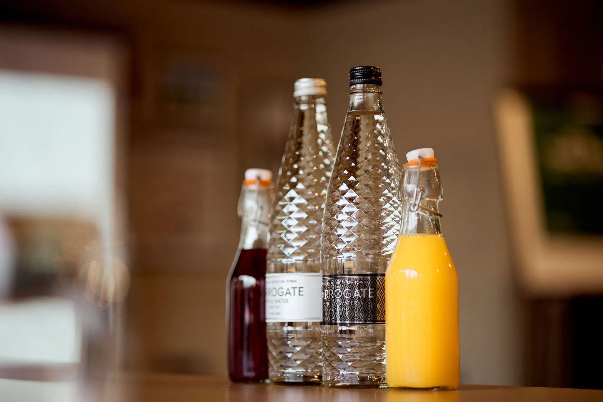 Selection of bottled drinks on a table, including spring water, orange juice and a dark cordial in a softly lit interior.