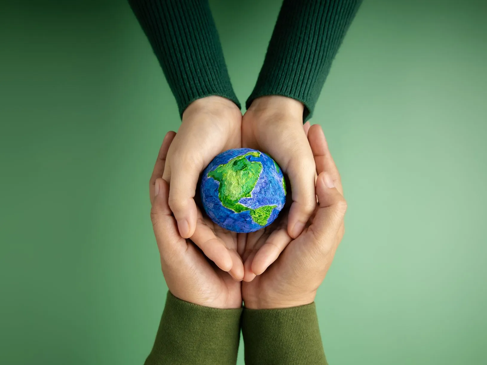 Two pairs of hands gently holding a small model of the Earth against a green background.