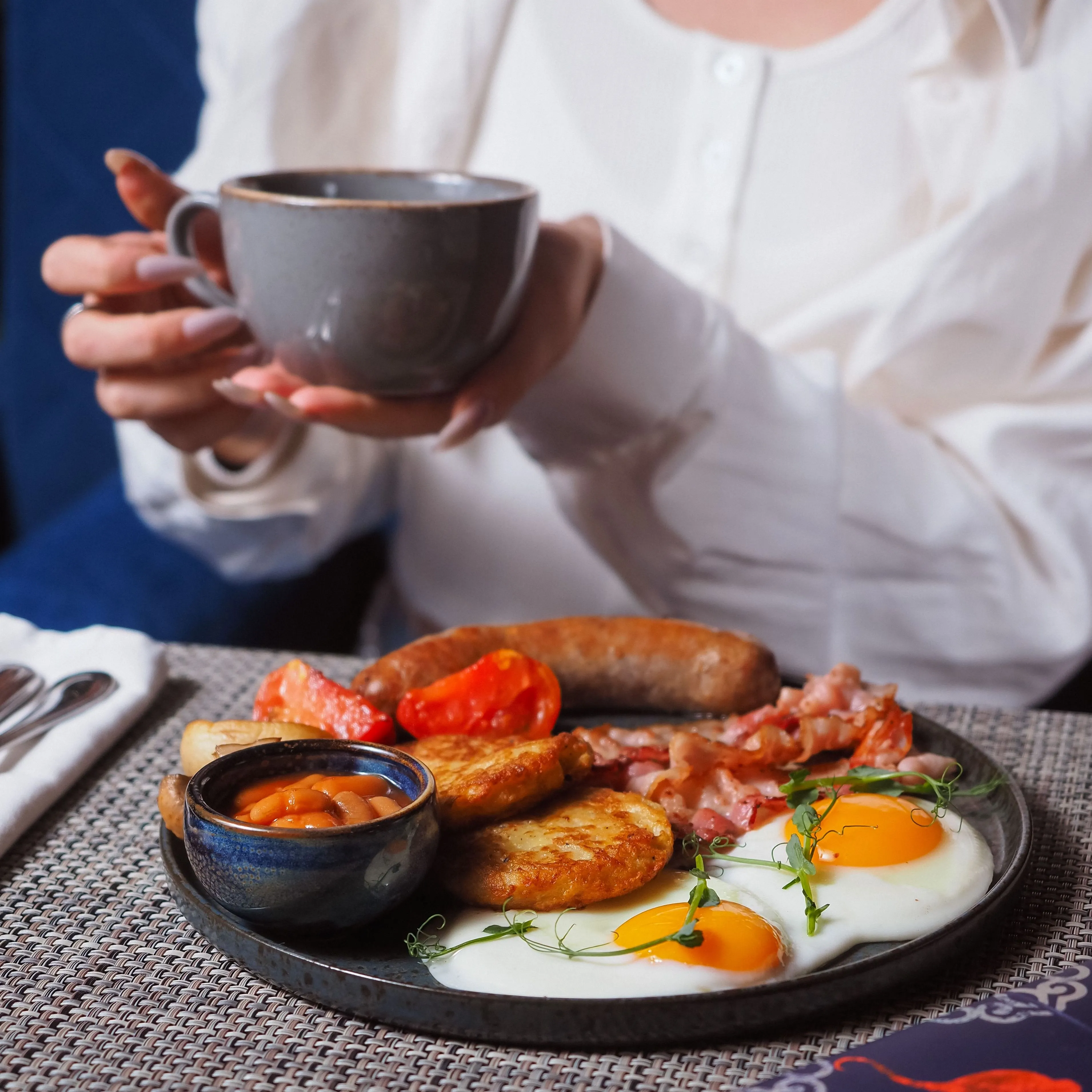 Woman holding a cup of coffee beside a full cooked breakfast with eggs, bacon, sausage, beans and hash browns