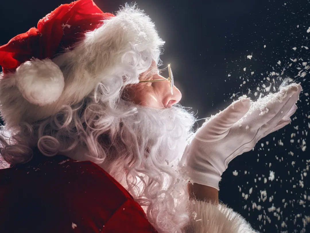 Father Christmas in a red suit and hat blows snowflakes from his gloved hand against a dark night sky.