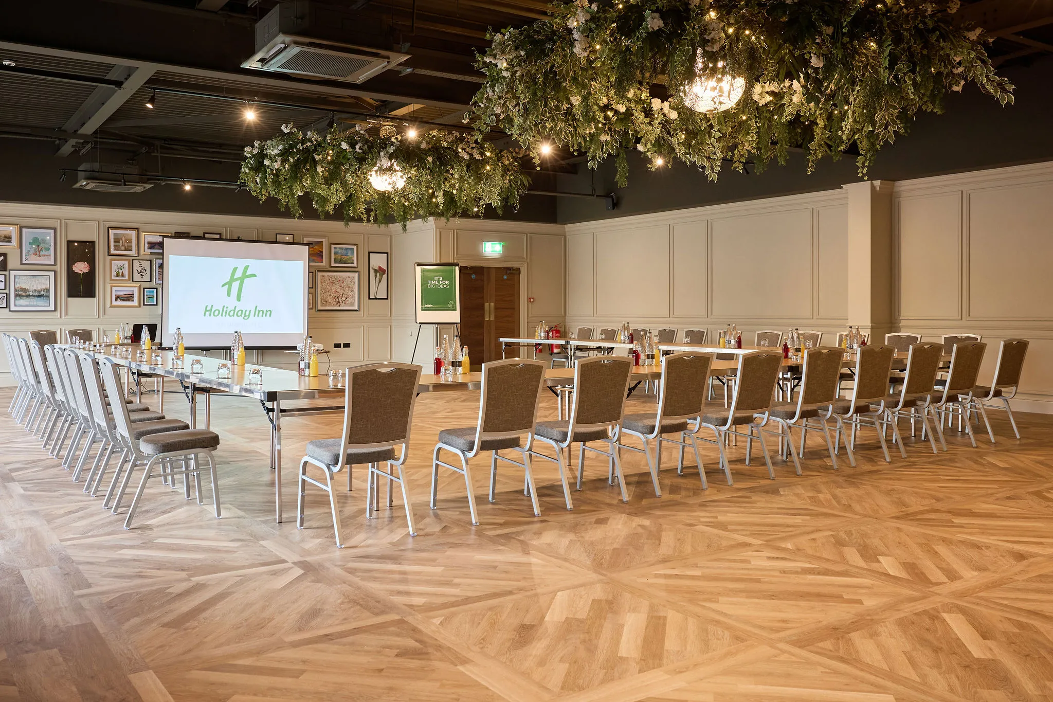 Conference room at Holiday Inn set up in a U-shape with tables, chairs and a projector screen beneath hanging floral installations