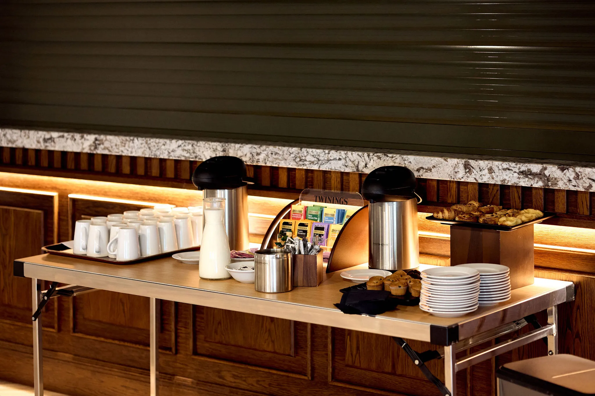 Refreshment table with tea, coffee, milk, mugs and pastries in a warmly lit interior
