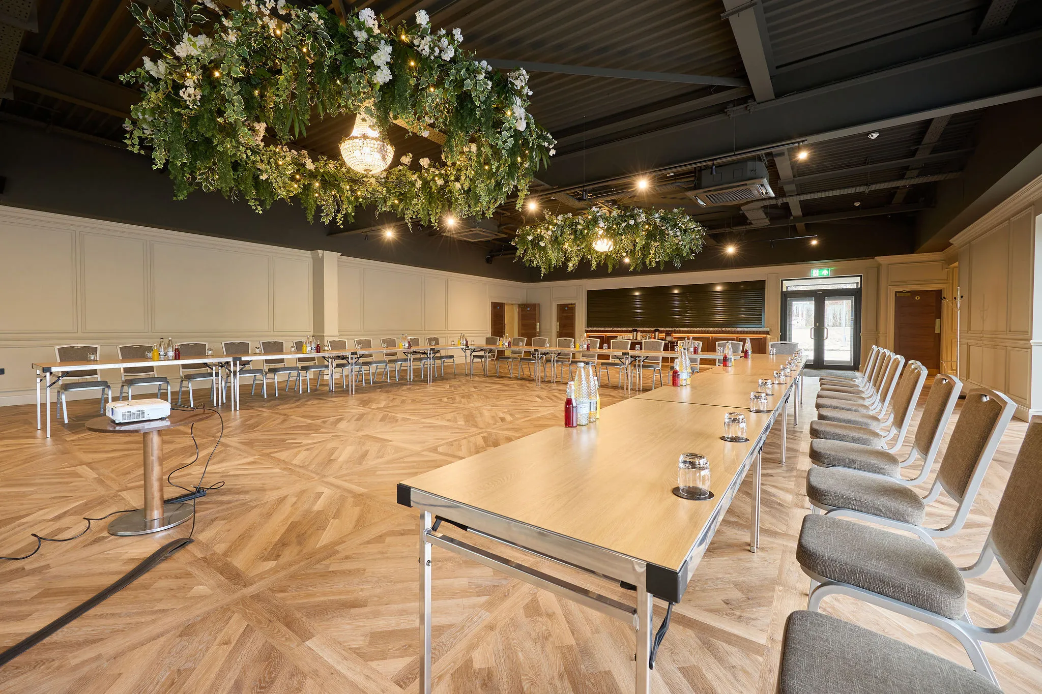Elegant function room set up in a U-shape conference layout with wooden tables, cushioned chairs, chandeliers and suspended floral greenery.