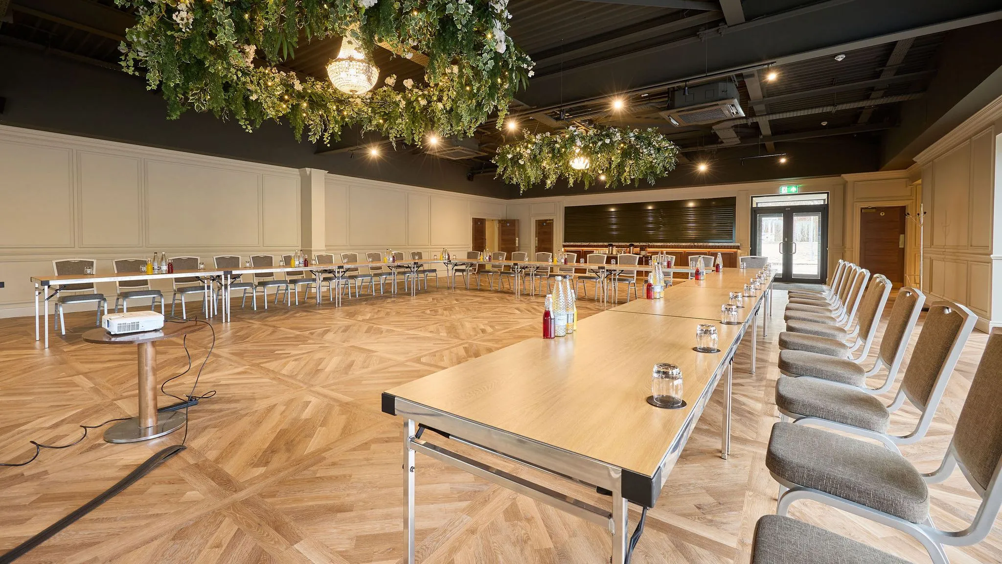 Elegant function room set up in a U-shape conference layout with wooden tables, cushioned chairs, chandeliers and suspended floral greenery.