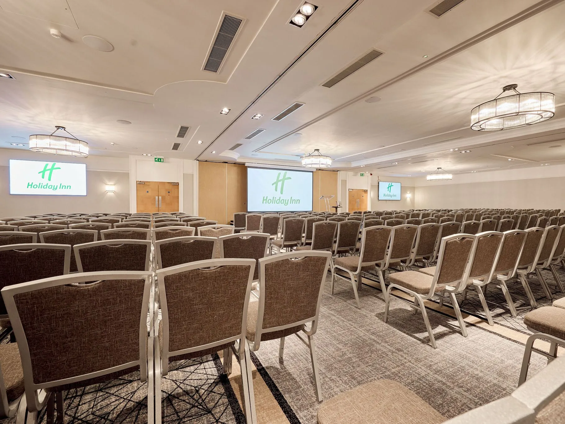 Large Holiday Inn conference room set up theatre-style with rows of chairs facing presentation screens and a lectern.