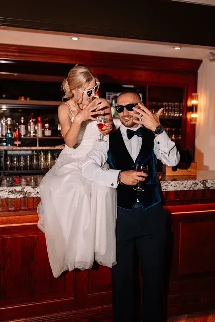Stylish wedding couple wearing sunglasses and holding drinks at a warmly lit bar