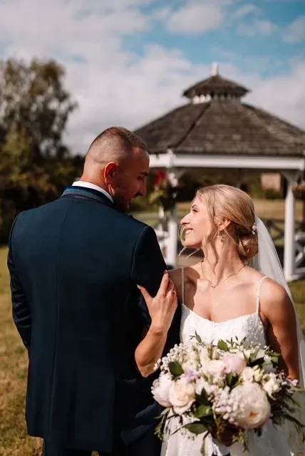 Bride holding a bouquet smiles at groom beside a gazebo during an outdoor wedding.