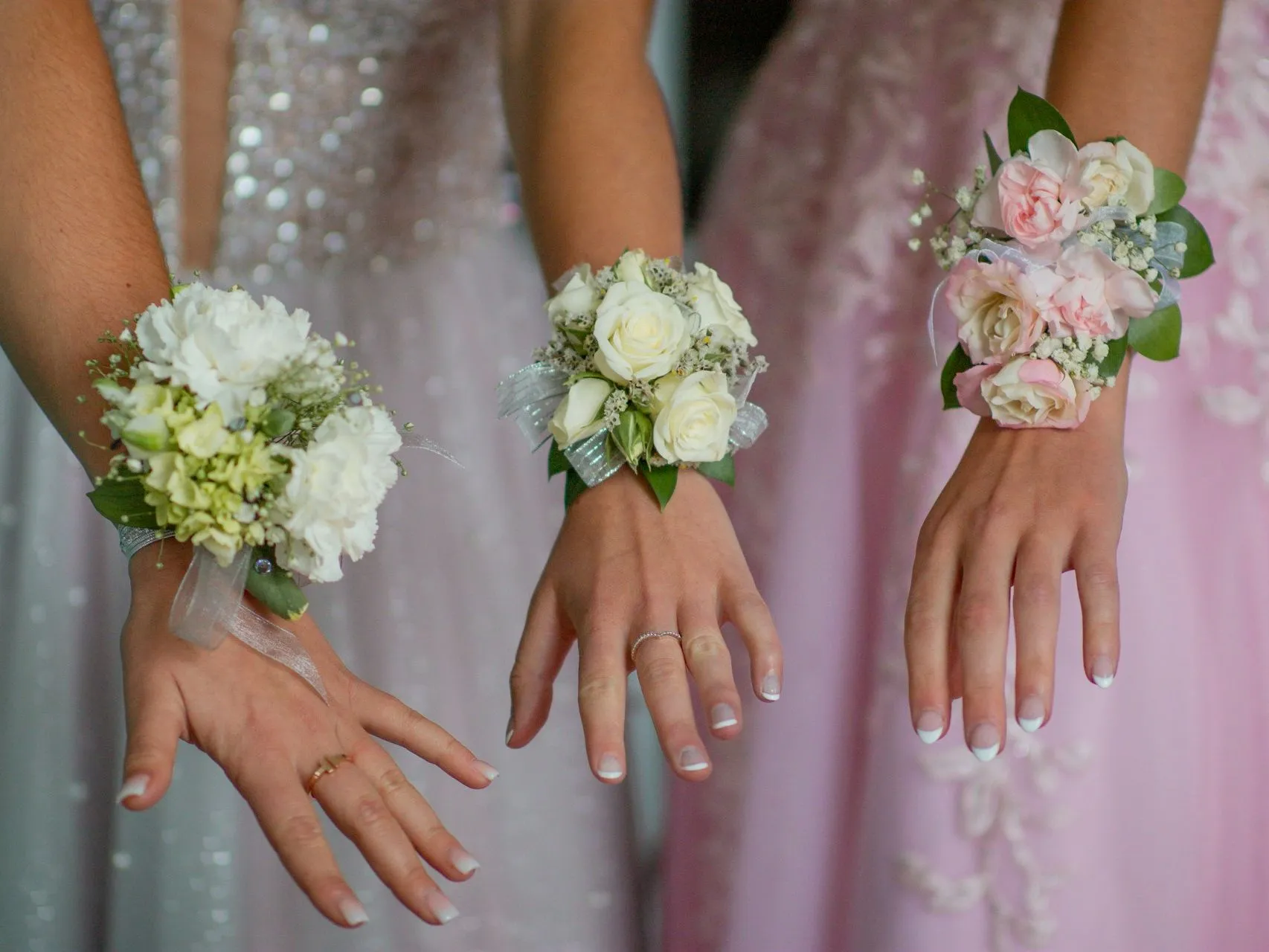 Three women in formal dresses wearing floral wrist corsages in white and blush pink.