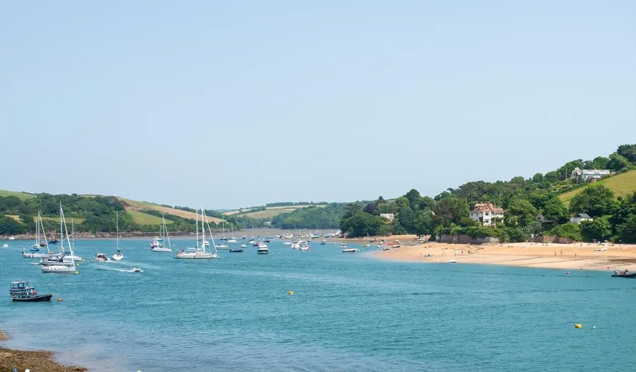 Sailboats moored in a calm estuary beside a sandy beach and green hills at Salcombe.