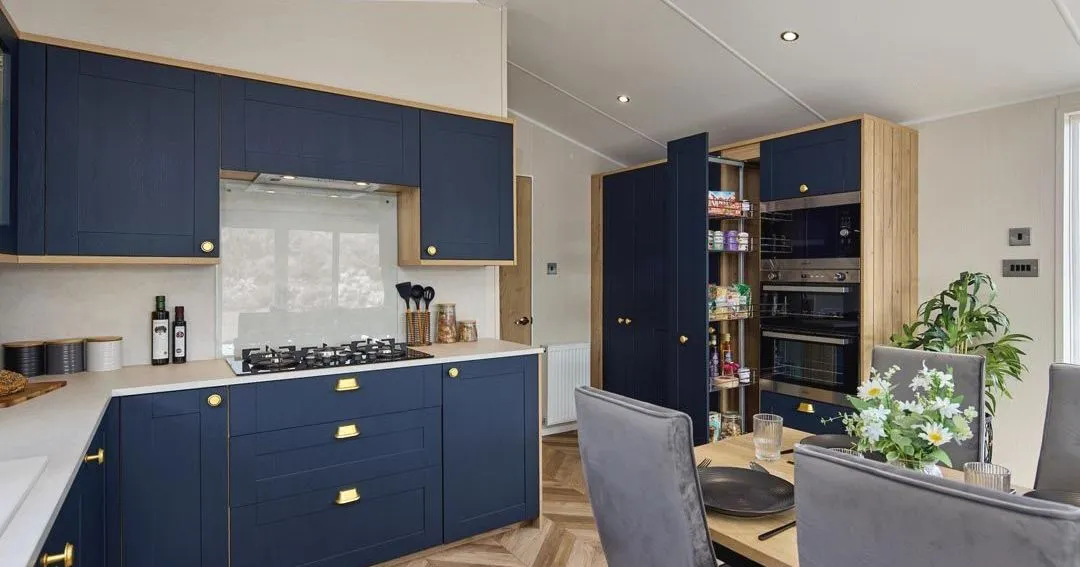 Modern kitchen with navy blue cabinets, brass handles and built-in ovens beside a dining table