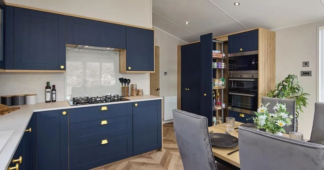 Modern kitchen with navy blue cabinets, brass handles and built-in ovens beside a dining table
