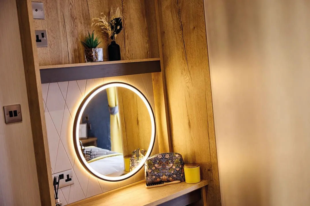 Round illuminated mirror above a wooden dressing table with toiletry bag and candle