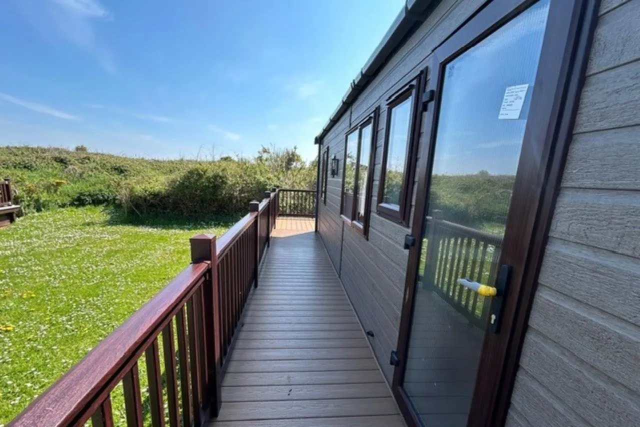Wooden decking walkway alongside a grey static caravan with railing and glass door, overlooking a grassy field