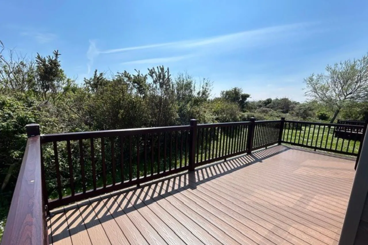 Timber deck with dark railing overlooking green shrubs and trees under a blue sky