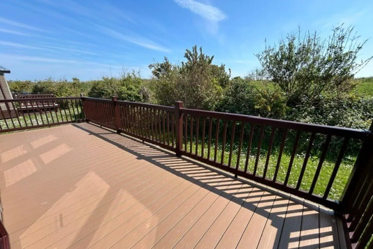 Sunlit wooden deck with dark railing overlooking green shrubs and trees