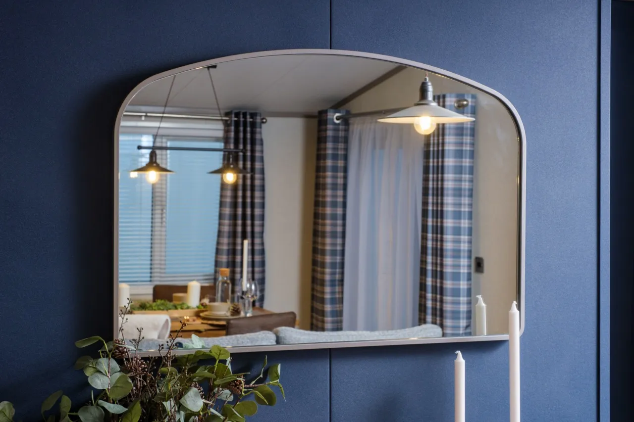 Rounded rectangular mirror on a dark blue wall reflecting a dining area with pendant lights and checked curtains