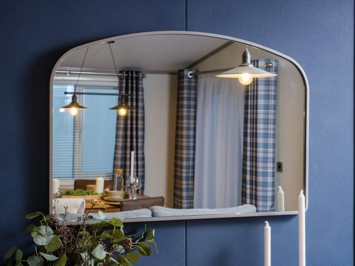 Rounded rectangular mirror on a dark blue wall reflecting a dining area with pendant lights and checked curtains
