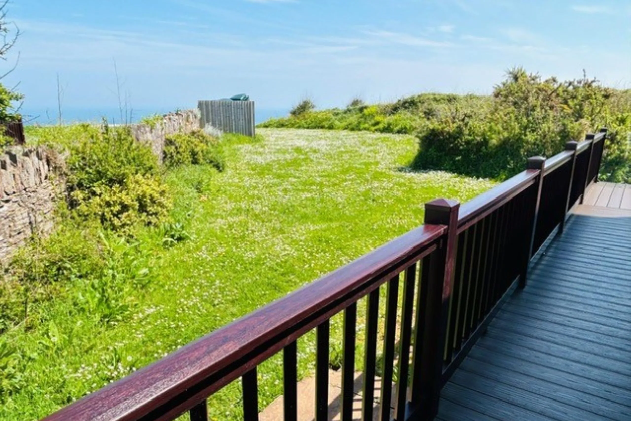 Wooden deck with dark railing overlooking a sunny grassy garden with coastal views