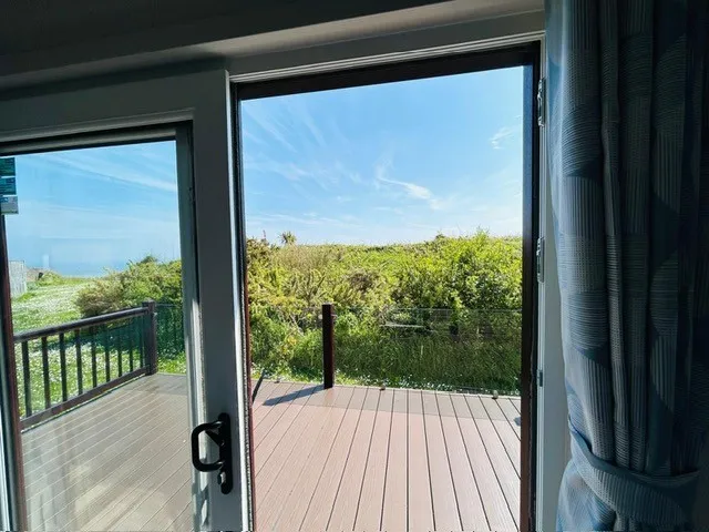 Open sliding glass door leading to a wooden deck with views of coastal greenery and blue sky