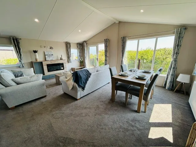Bright open-plan lounge and dining area with grey sofas, dining table and large windows overlooking greenery