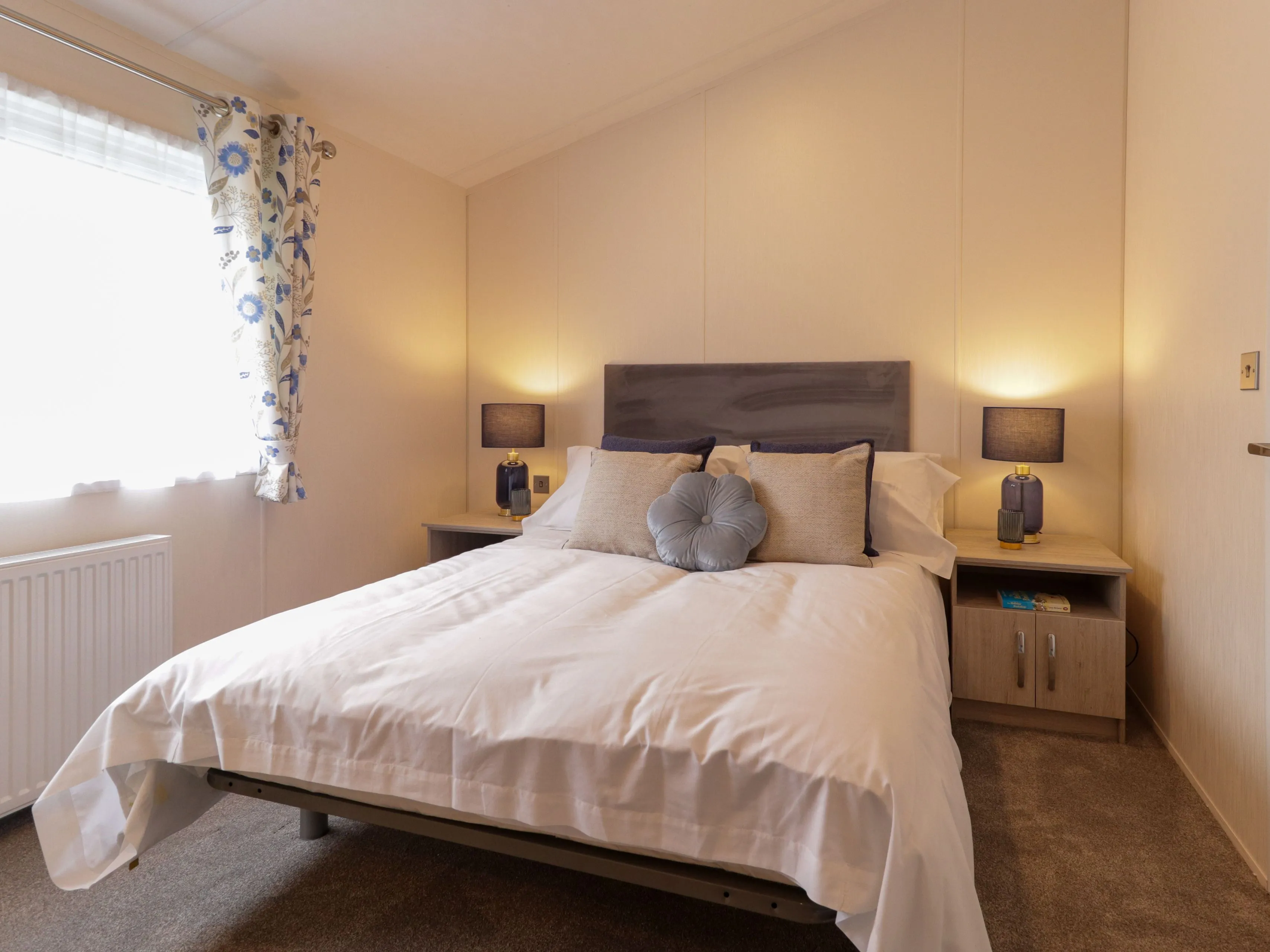 Cosy bedroom with double bed, grey headboard, bedside tables and lamps, and a window with floral curtains