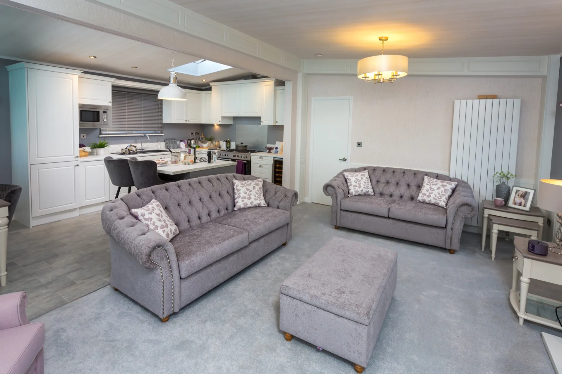 Open-plan living room and kitchen with grey tufted sofas, ottoman, and white cabinetry