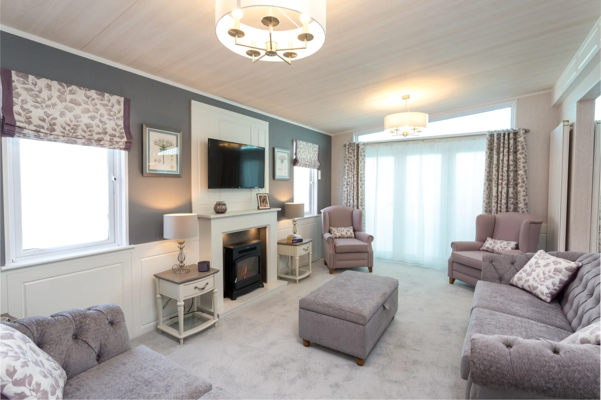 Bright modern living room with grey walls, wall-mounted TV above a fireplace, armchairs, sofas, ottoman and large patio doors with patterned curtains.