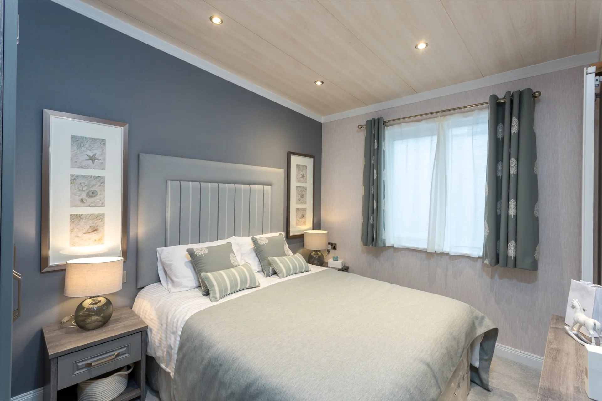 Modern bedroom with grey upholstered headboard, bedside lamps, framed wall art, and a window with patterned curtains