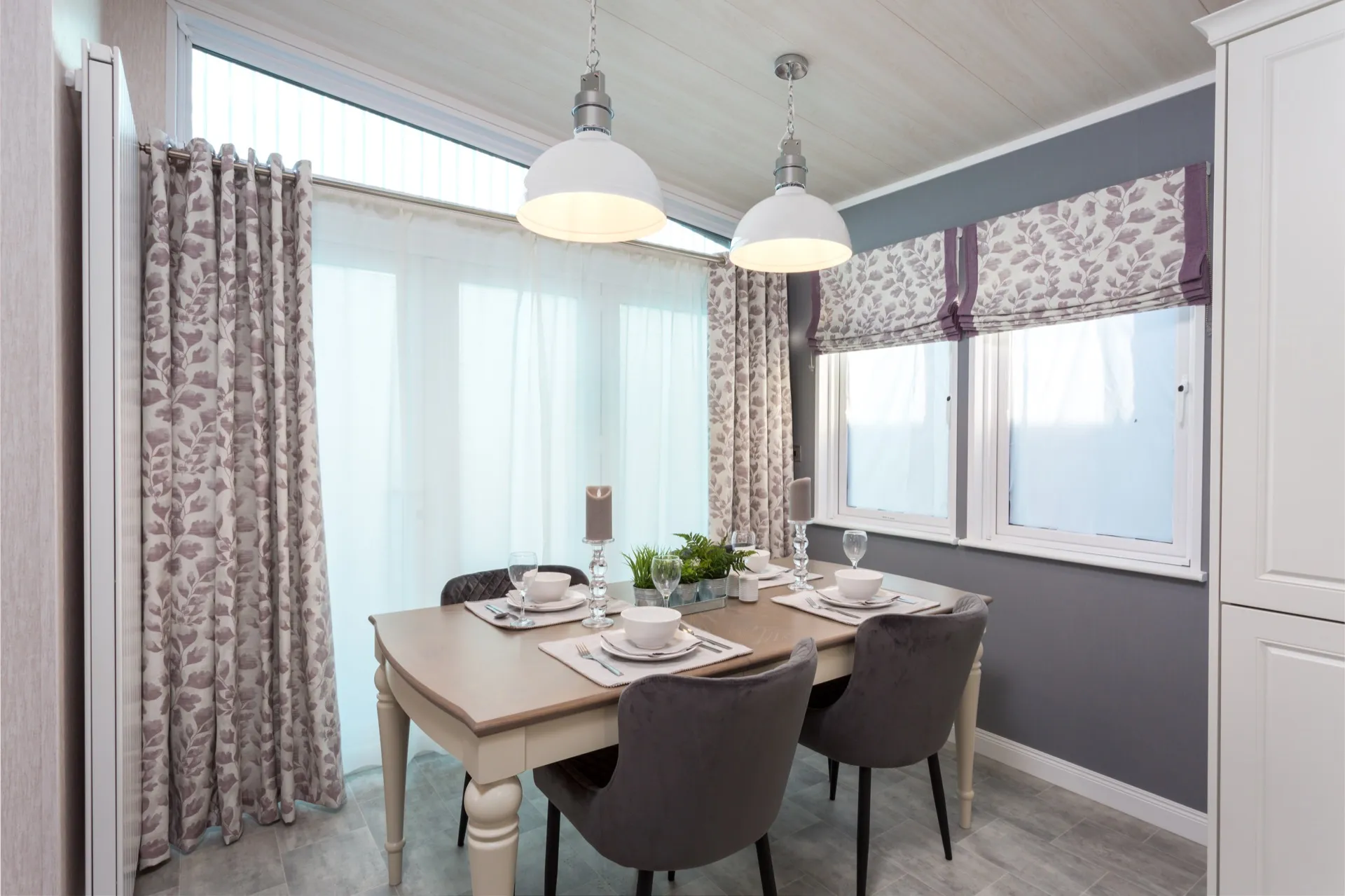 Modern dining area with four place settings, grey upholstered chairs, and floral curtains under white pendant lights