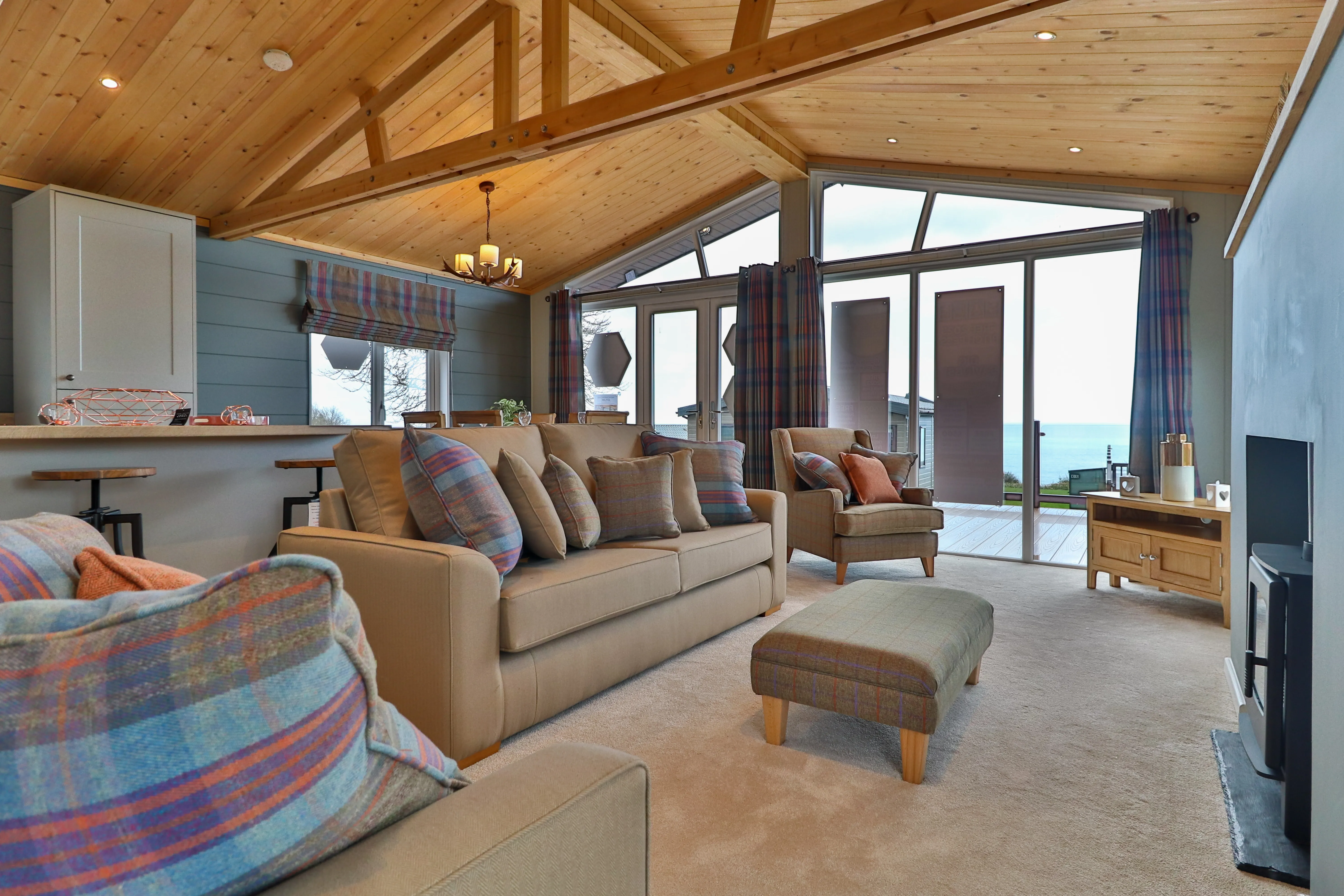 Cosy living room with vaulted wooden ceiling, plaid cushions, and large glass doors opening to a sea view
