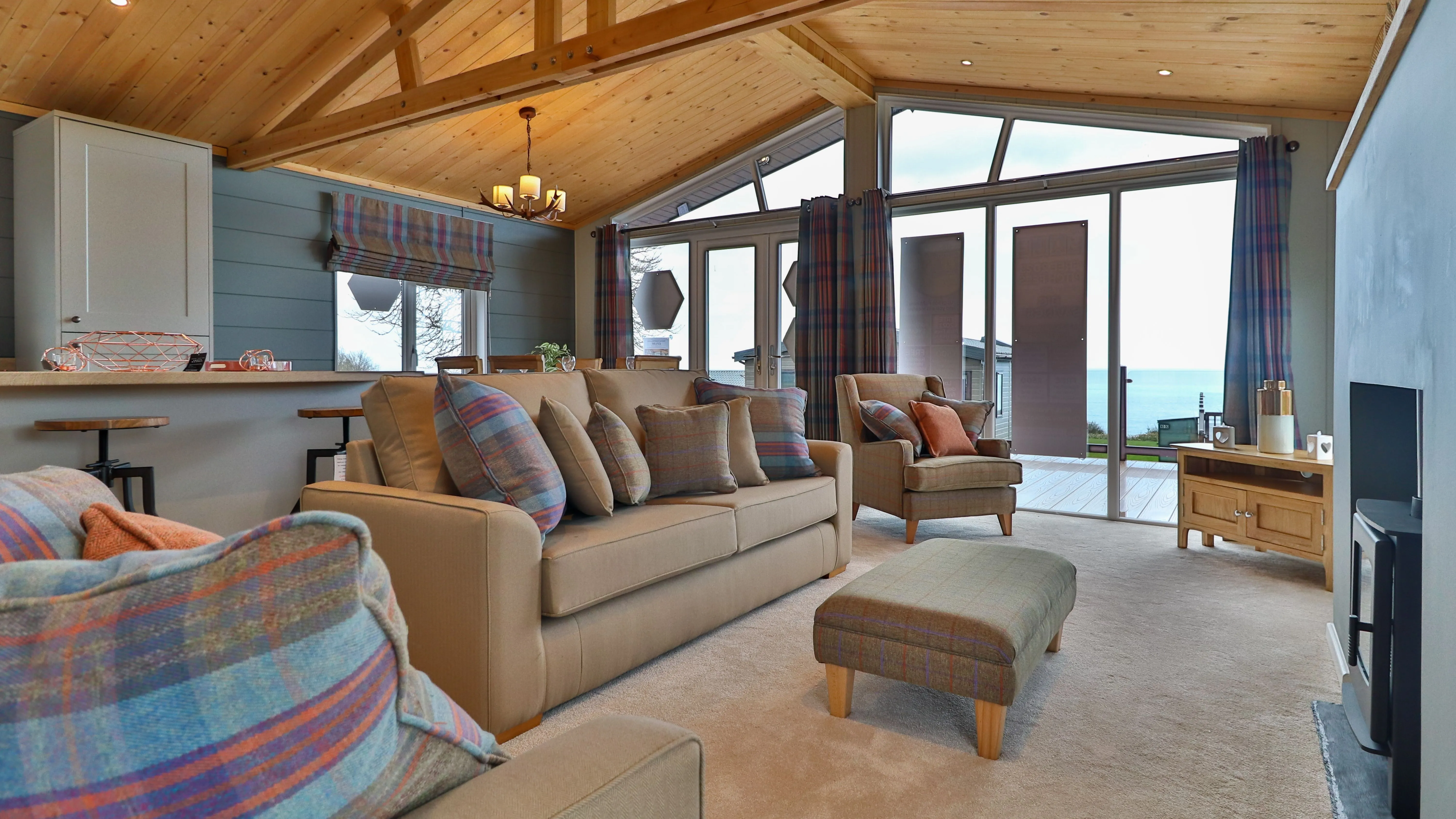 Cosy living room with vaulted wooden ceiling, plaid cushions, and large glass doors opening to a sea view