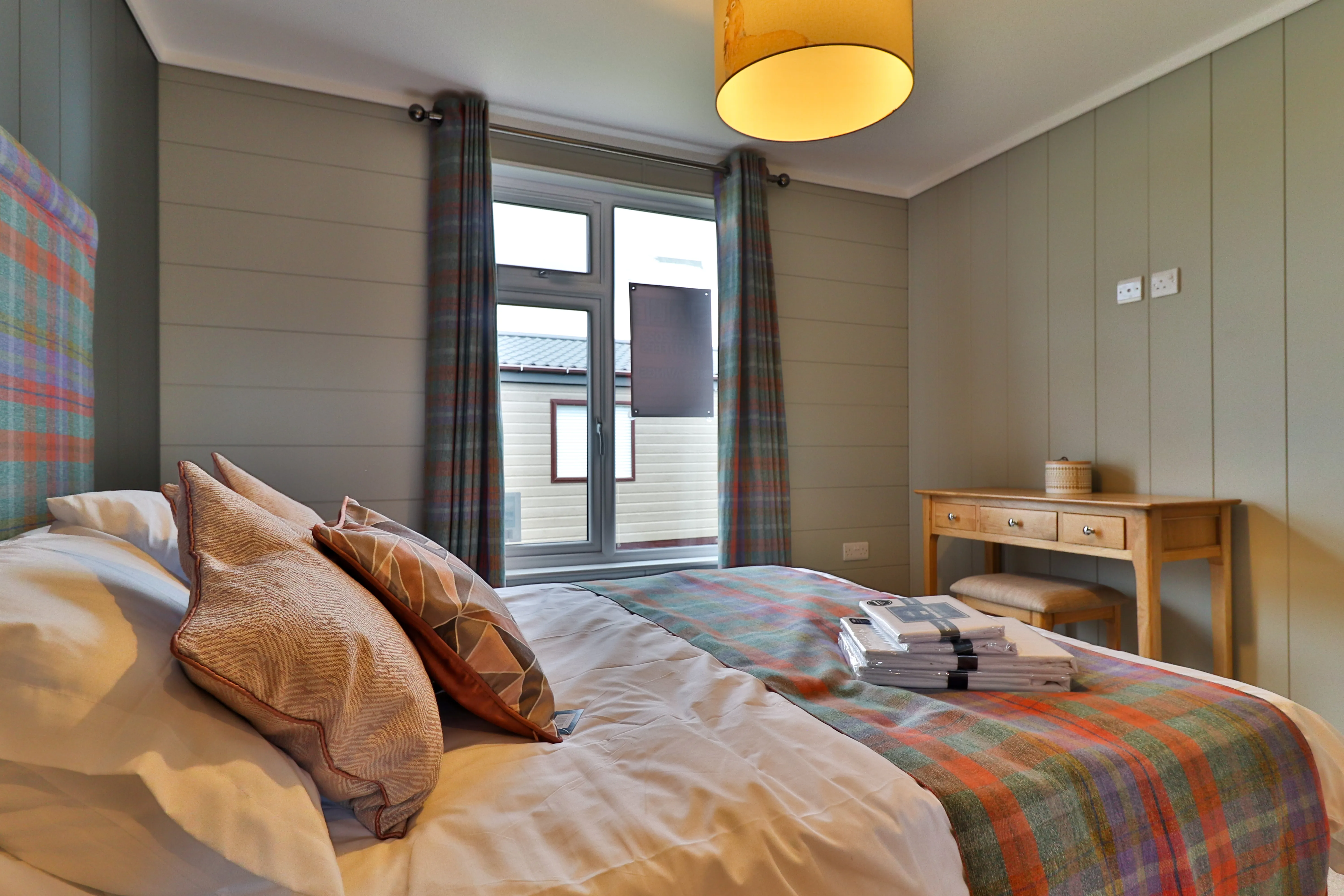 Cosy bedroom with a neatly made bed, plaid throw and curtains, and a wooden dressing table by the window