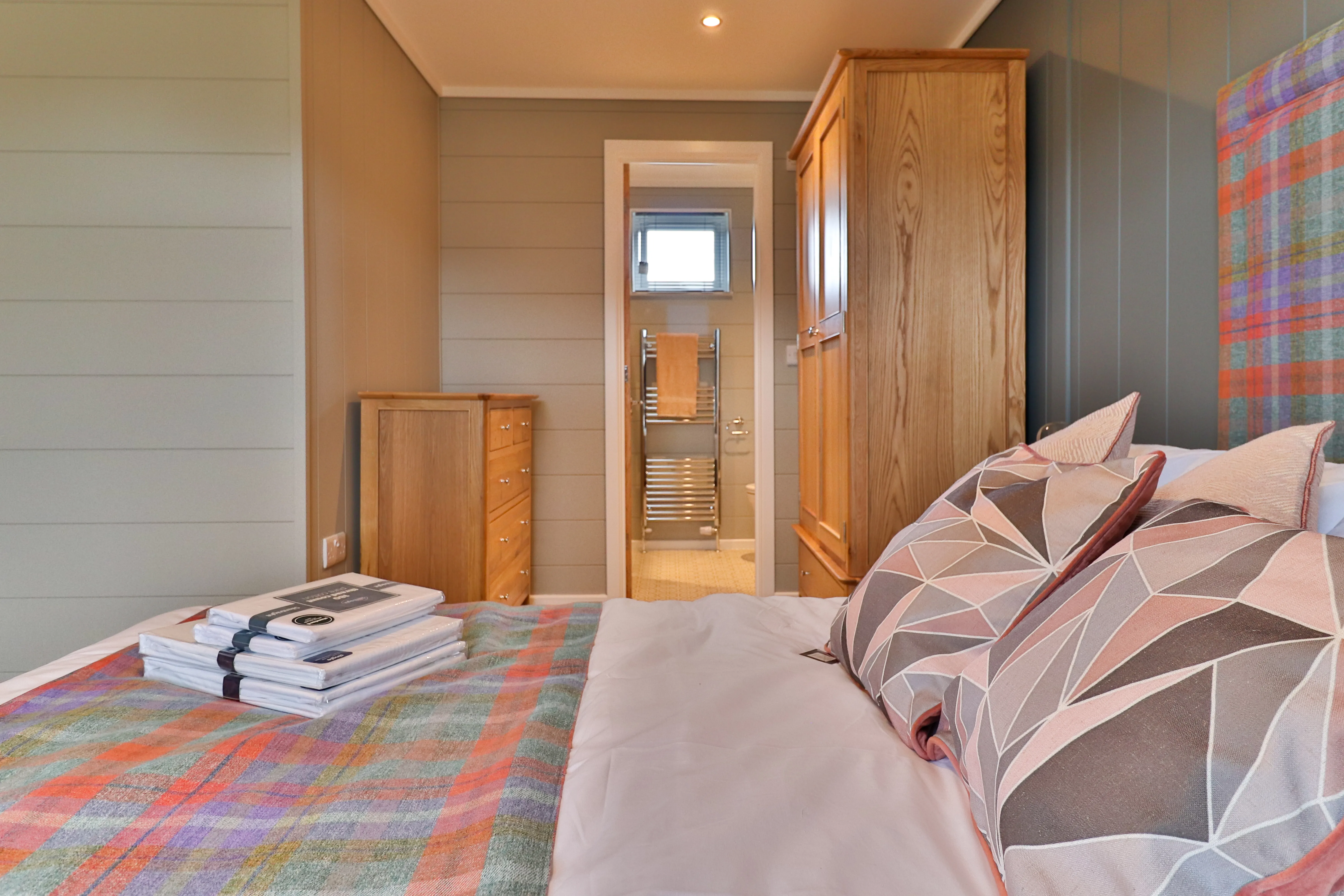 Cosy bedroom with double bed, patterned cushions and wooden wardrobe, looking through to an en-suite bathroom