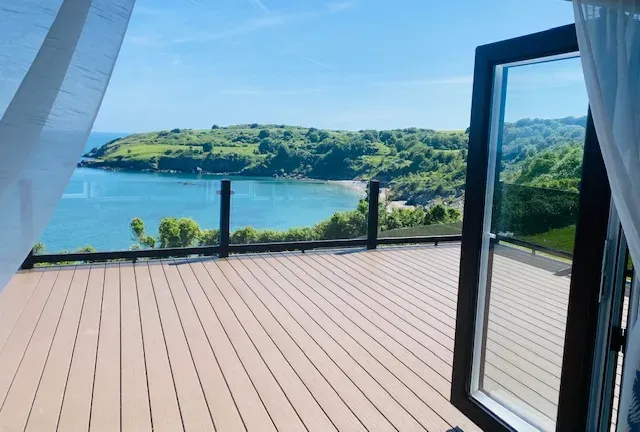 Sunny deck with open glass door overlooking a calm bay and green hills