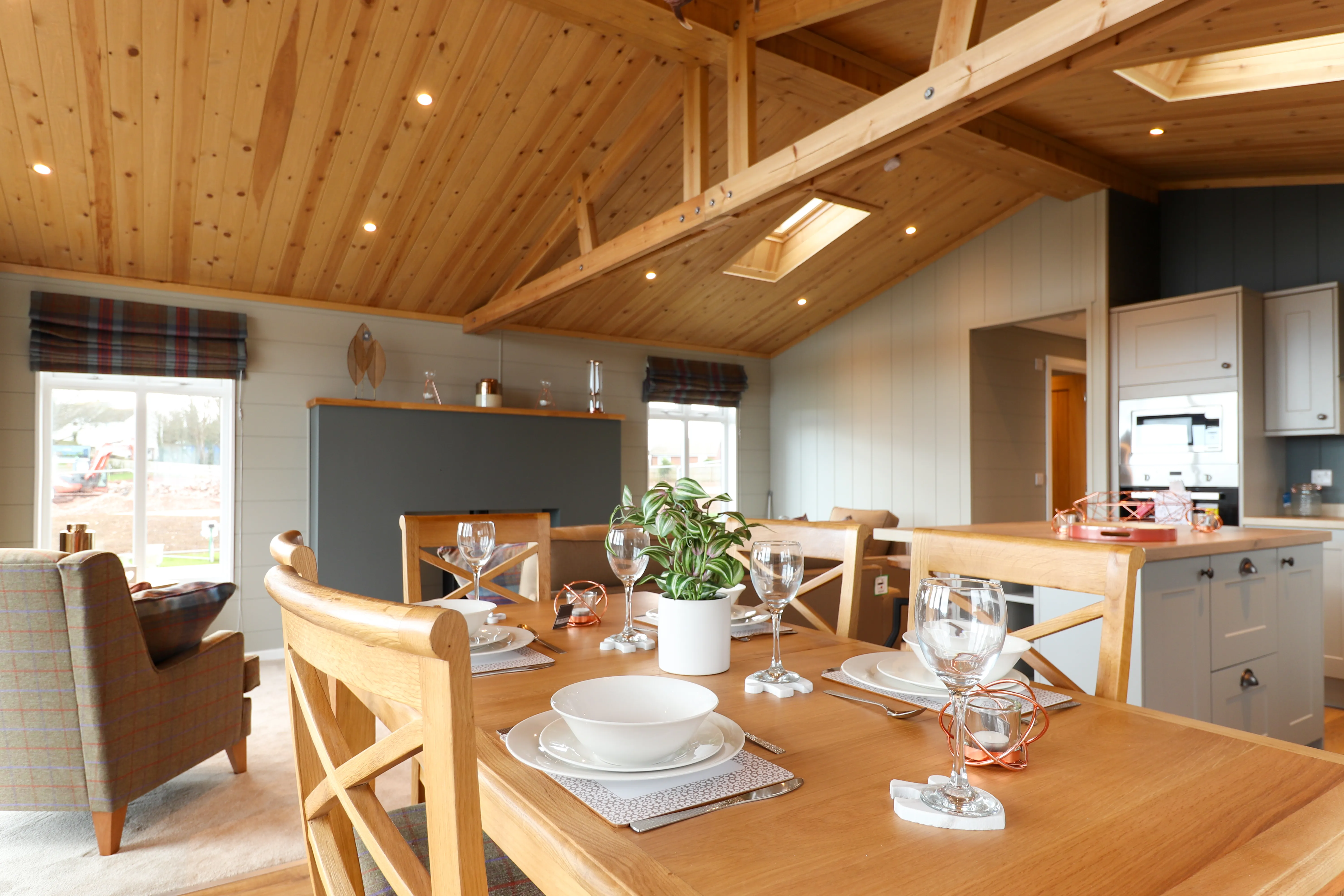 Open-plan dining area with wooden table set for four beneath a vaulted timber ceiling, adjoining a modern kitchen.