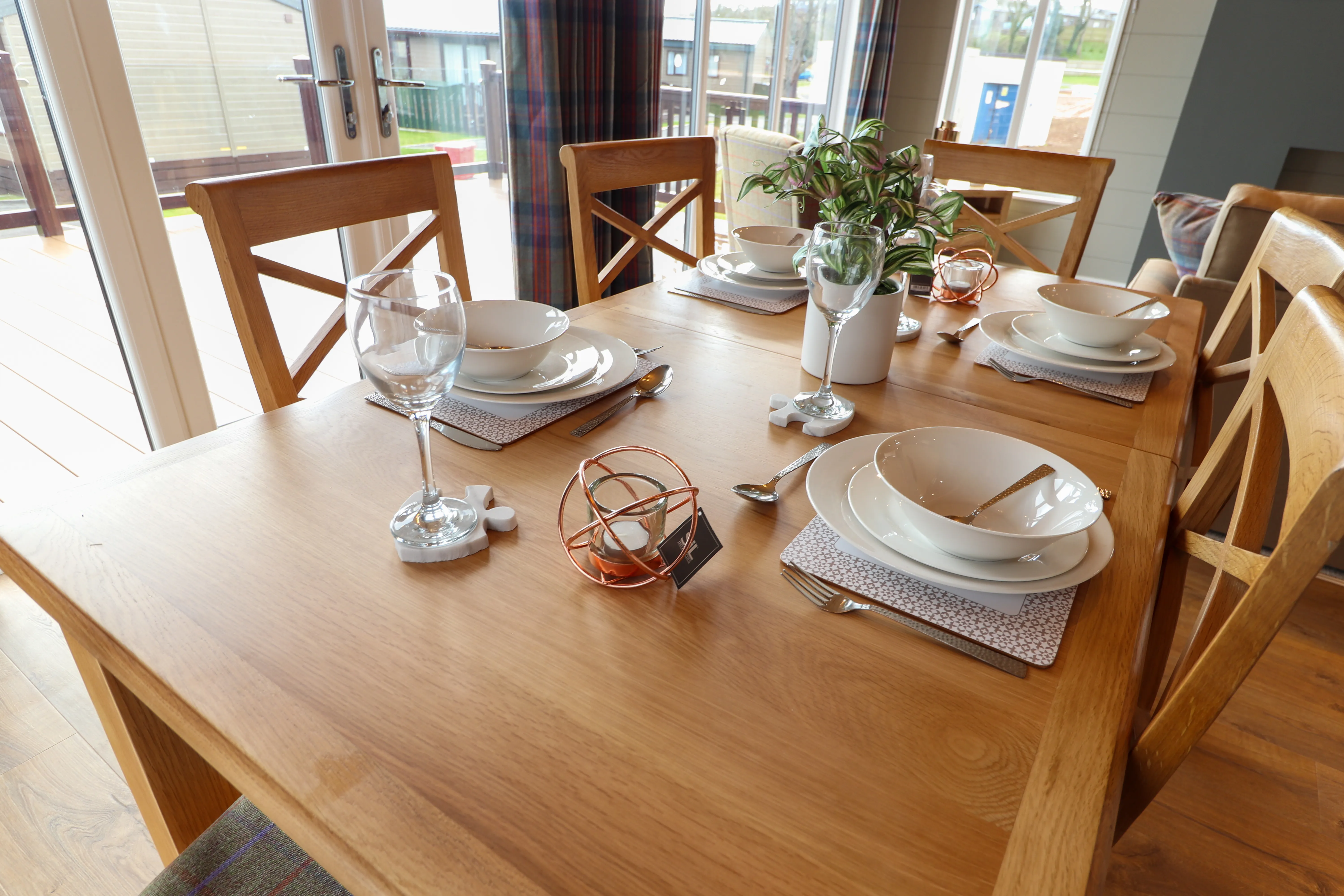Wooden dining table set with bowls, plates, cutlery and wine glasses by a bright window