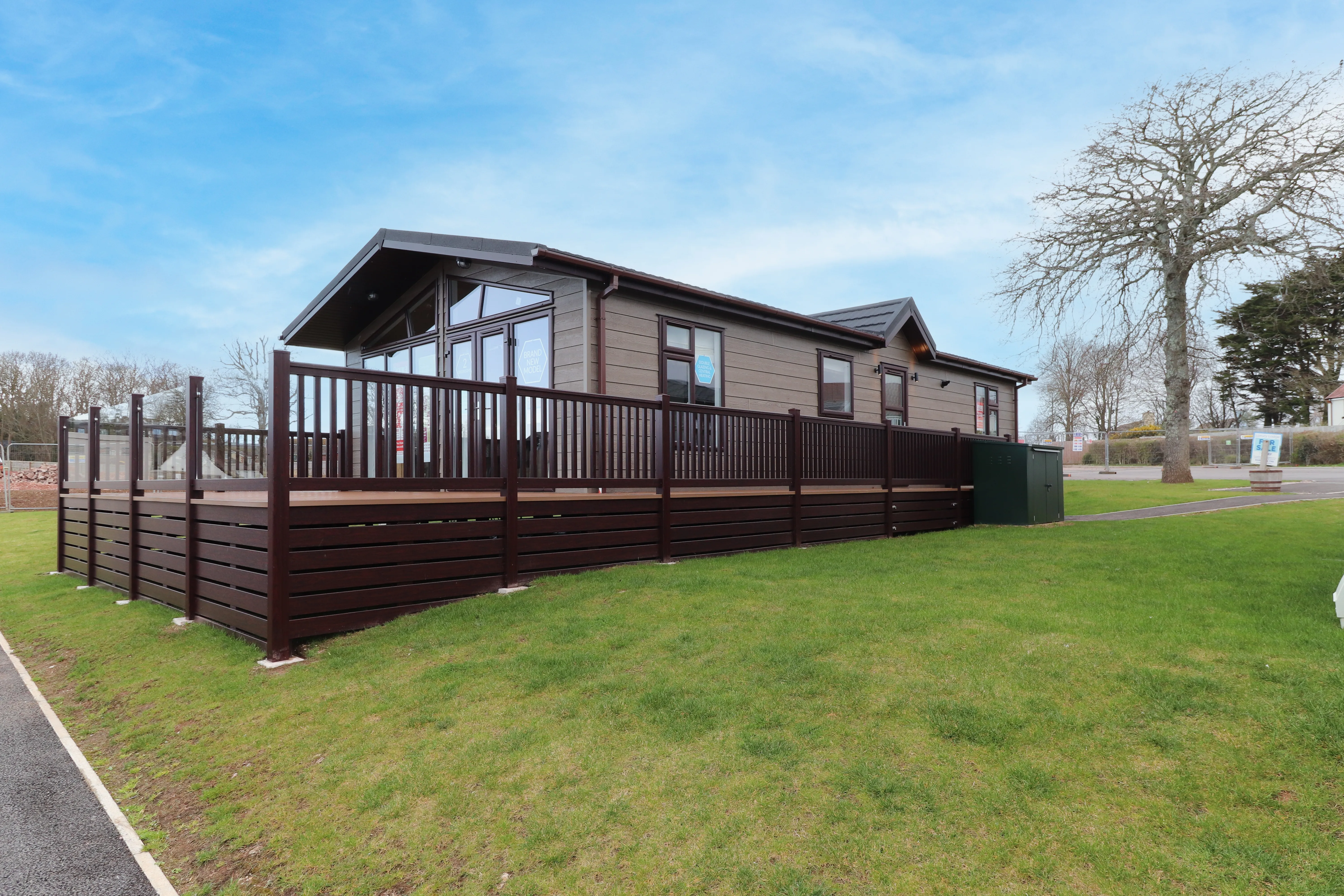Modern lodge-style caravan with a large wooden deck on a grassy plot under a blue sky