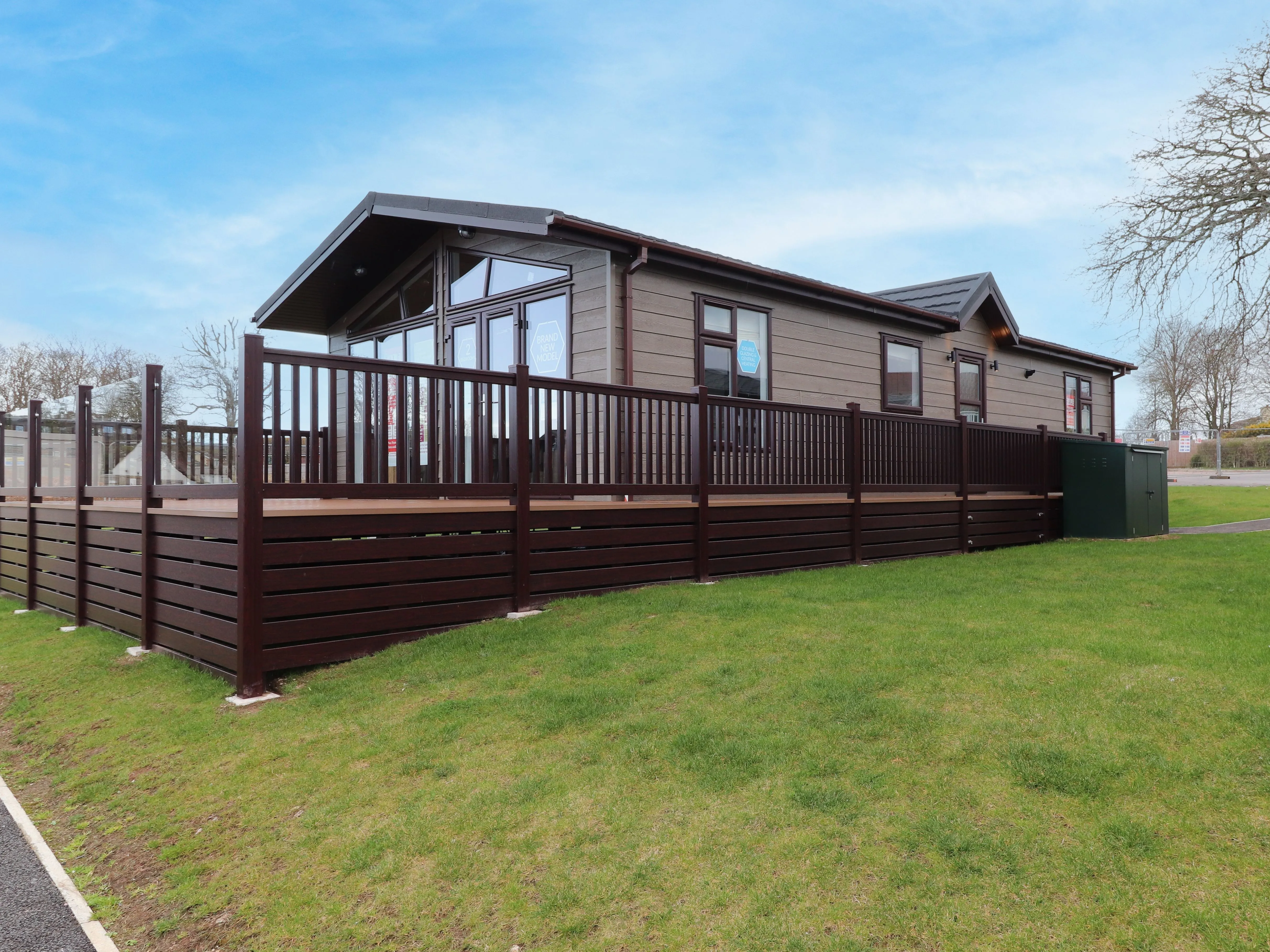 Modern lodge-style caravan with a large wooden deck on a grassy plot under a blue sky