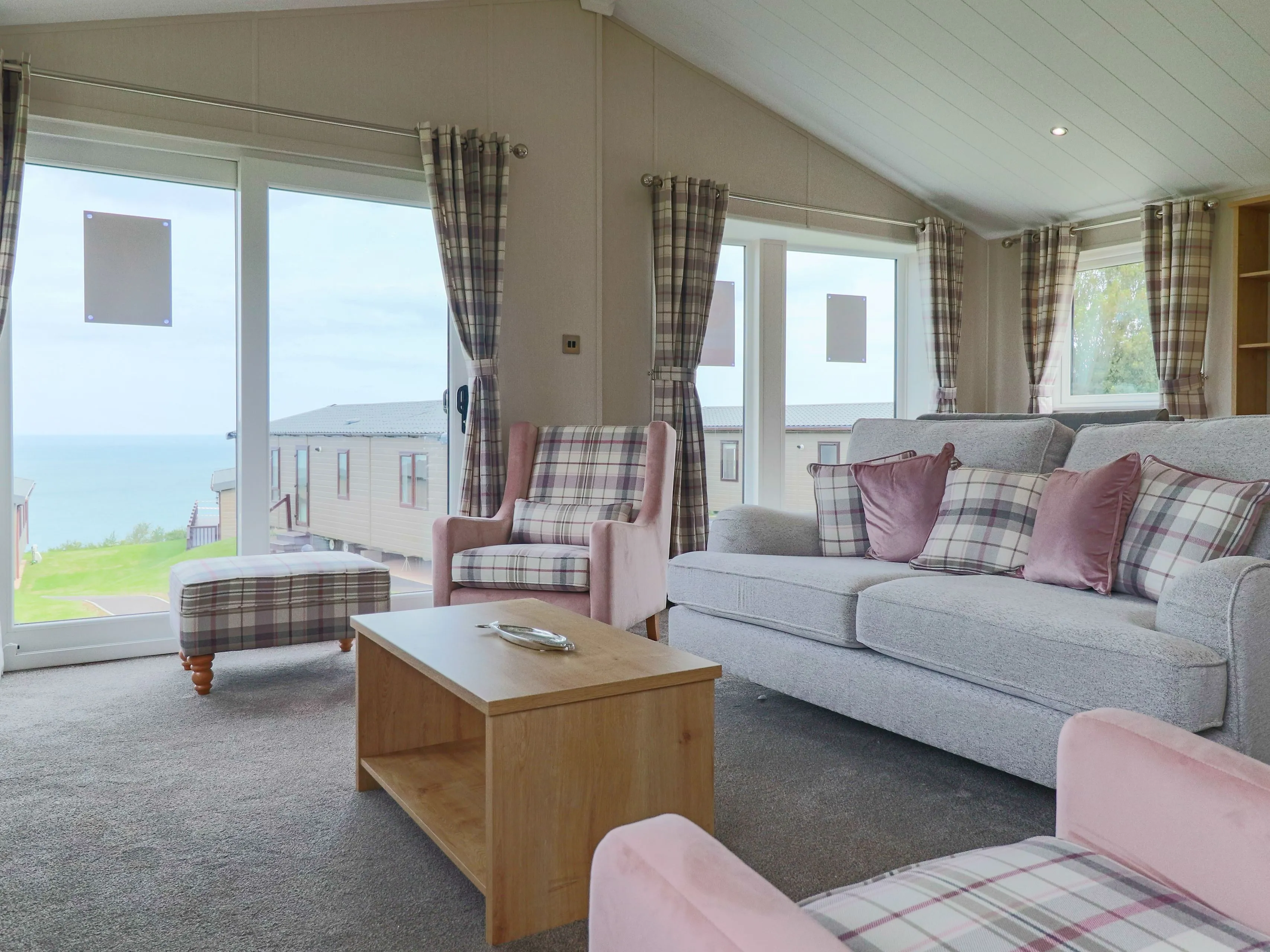 Bright lounge with grey sofa, pink armchairs and plaid curtains beside large windows overlooking the sea