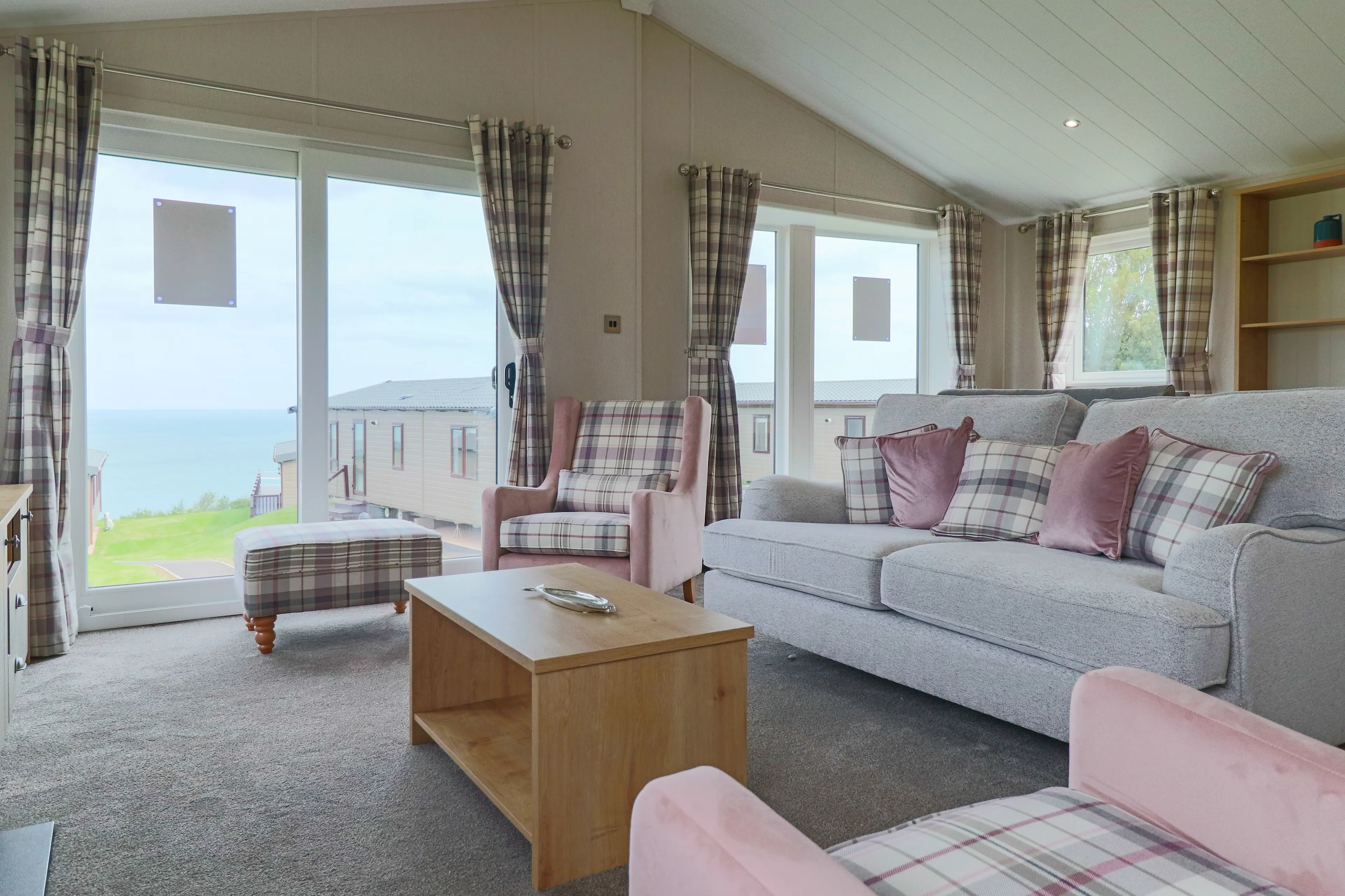 Bright lounge with grey sofa, pink armchairs and plaid curtains beside large windows overlooking the sea